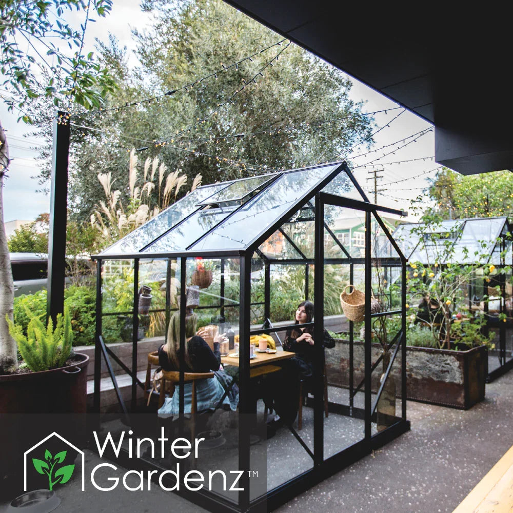 Two people sitting and chatting inside a small glass greenhouse patio pod outdoors