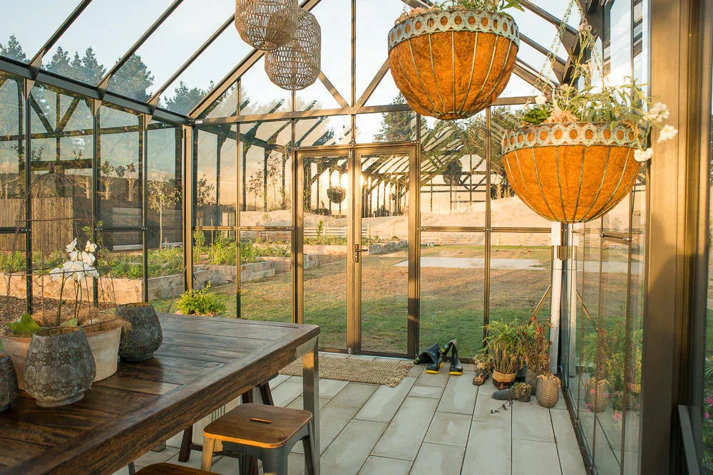 Interior of a spacious 12x16ft glass greenhouse with wooden table, hanging planters, garden tools, and sunlight