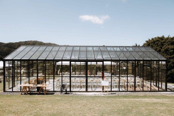 Glass greenhouse wedding venue with rows of white chairs and wooden tables in a scenic outdoor setting