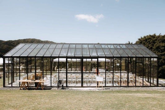 Glass greenhouse wedding venue with rows of white chairs and wooden tables in a scenic outdoor setting