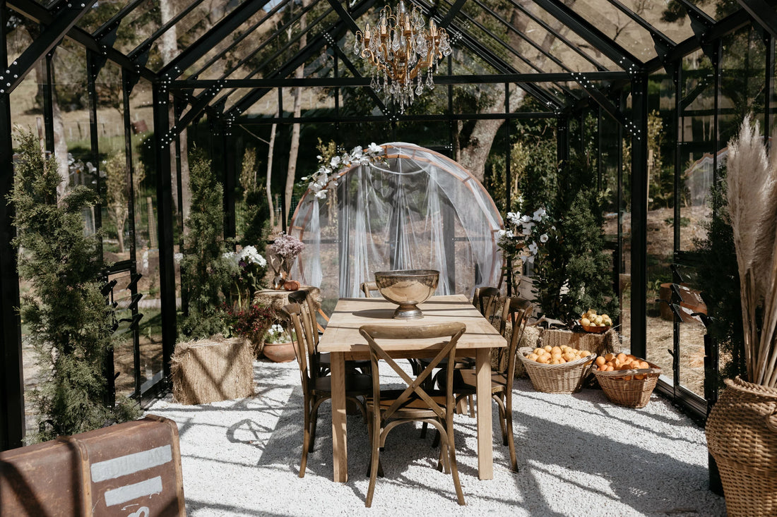 Rustic glass greenhouse dining space with wooden table, chairs, chandelier, flowers, and baskets of fruit