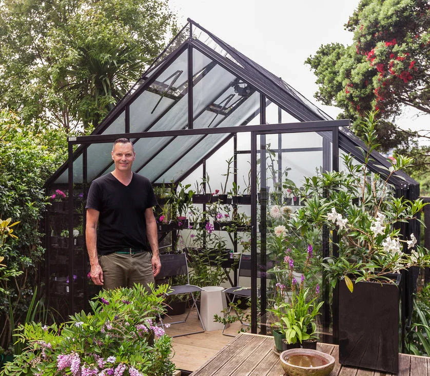Man standing in doorway of modern glass greenhouse filled with flowering plants and greenery