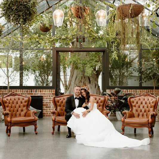 Bride and groom sitting on vintage leather sofa in a greenhouse wedding venue with hanging plants