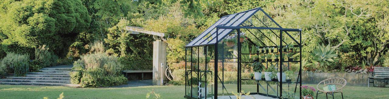 Modern glass greenhouse with potted plants in a lush home garden with trees and seating