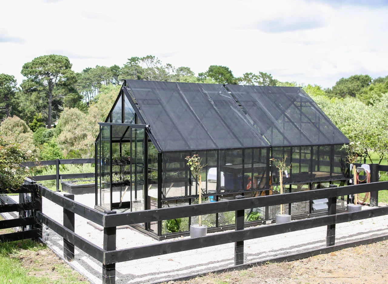Glasshouse greenhouse with black shade cloth covering, black fence, and small plants outside in a garden setting